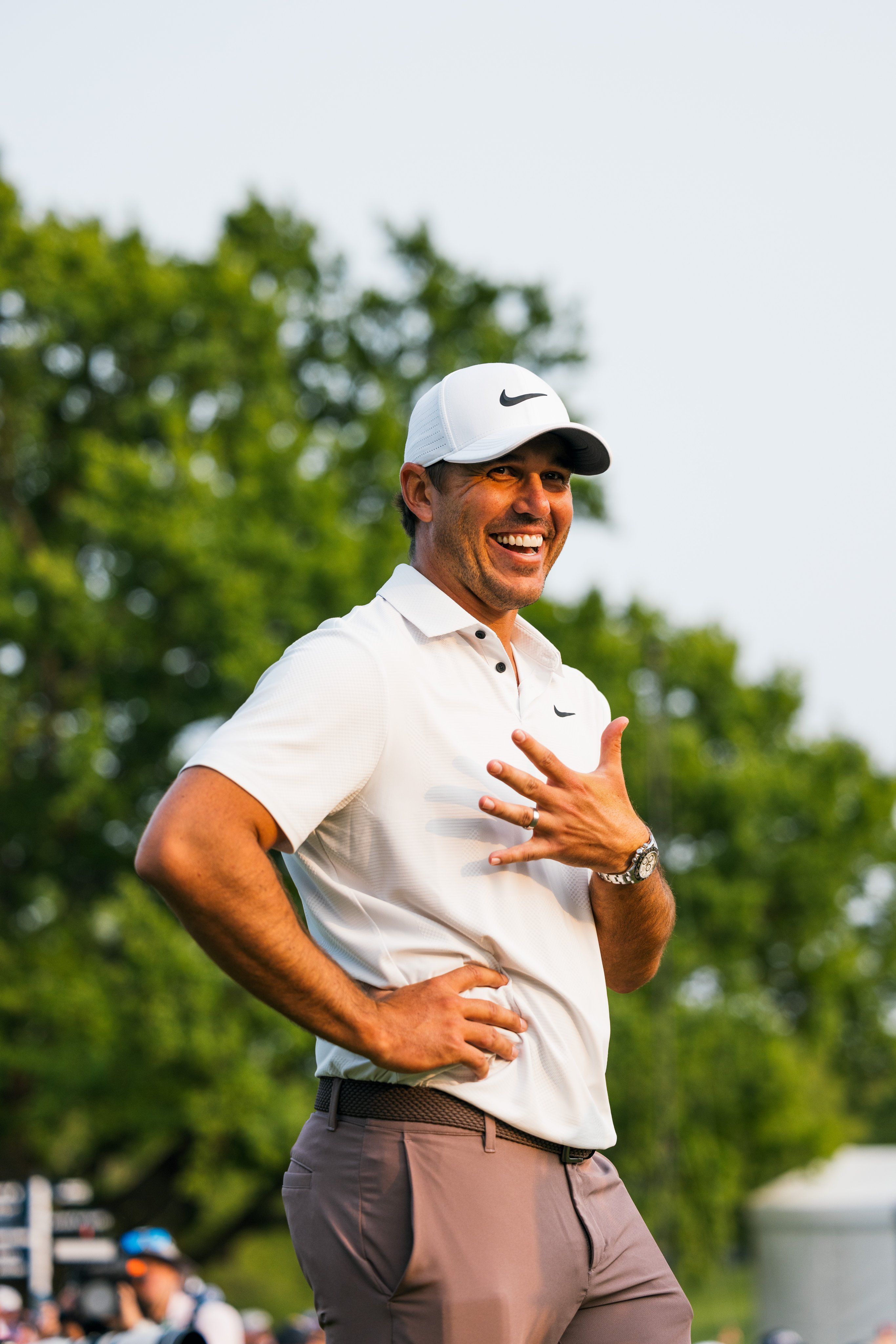 Golfer Brooks Koepka wearing a white golf shirt and brown golf pants with Nike logo is standing and laughing