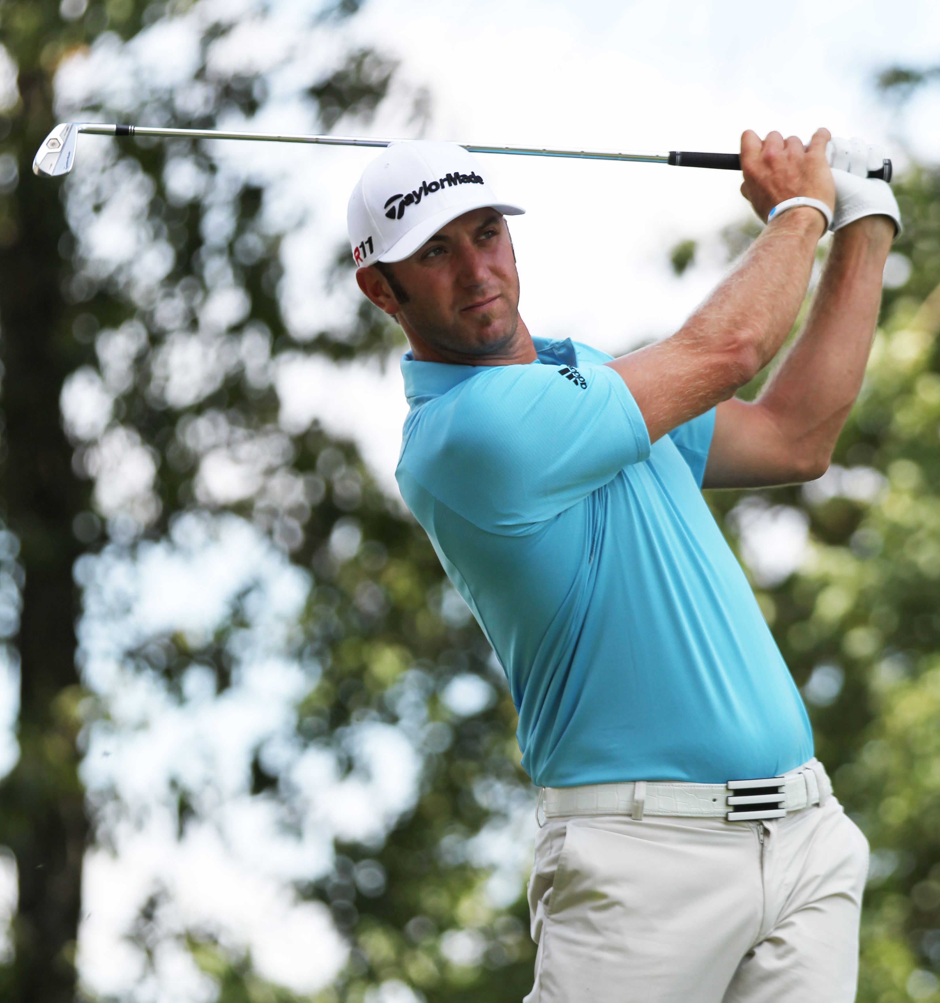 Golfer Dustin Johnson is wearing a blue shirt and off-white golf pants, watches his golf ball after hitting an iron shot.