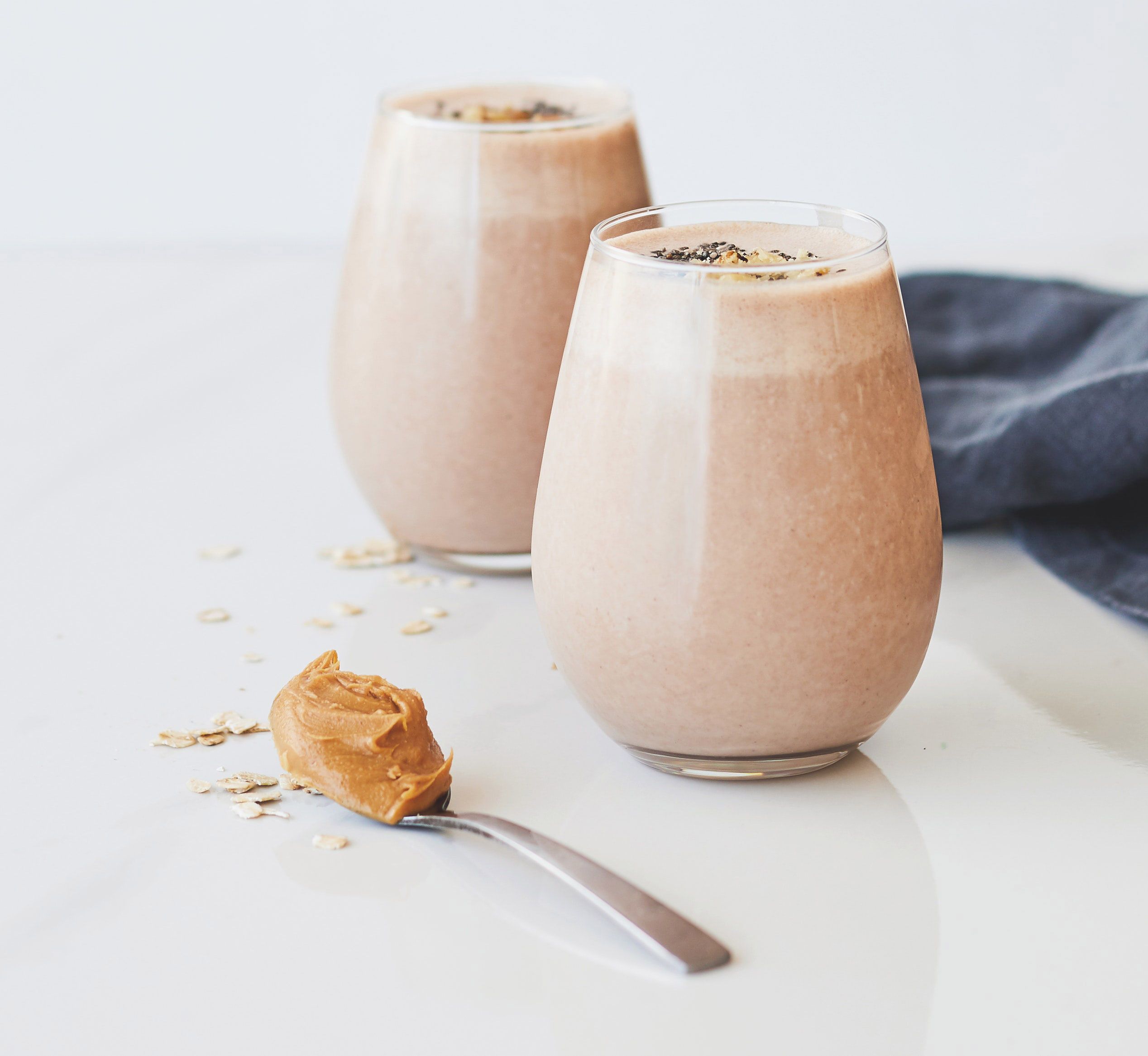 Two Glasses of Peanut Butter Oatmeal Smoothie on a counter with a spoon filled with peanut butter resting beside